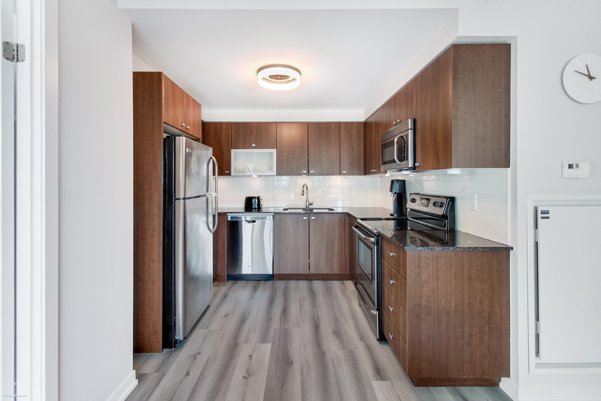 4 | Liberty Village Toronto | Your Online Neighbourhood Guide Modern U-shaped kitchen with dark wood cabinets, stainless steel fridge, stove, microwave, and a white tile backsplash under a bright ceiling light, gray wood floors.
