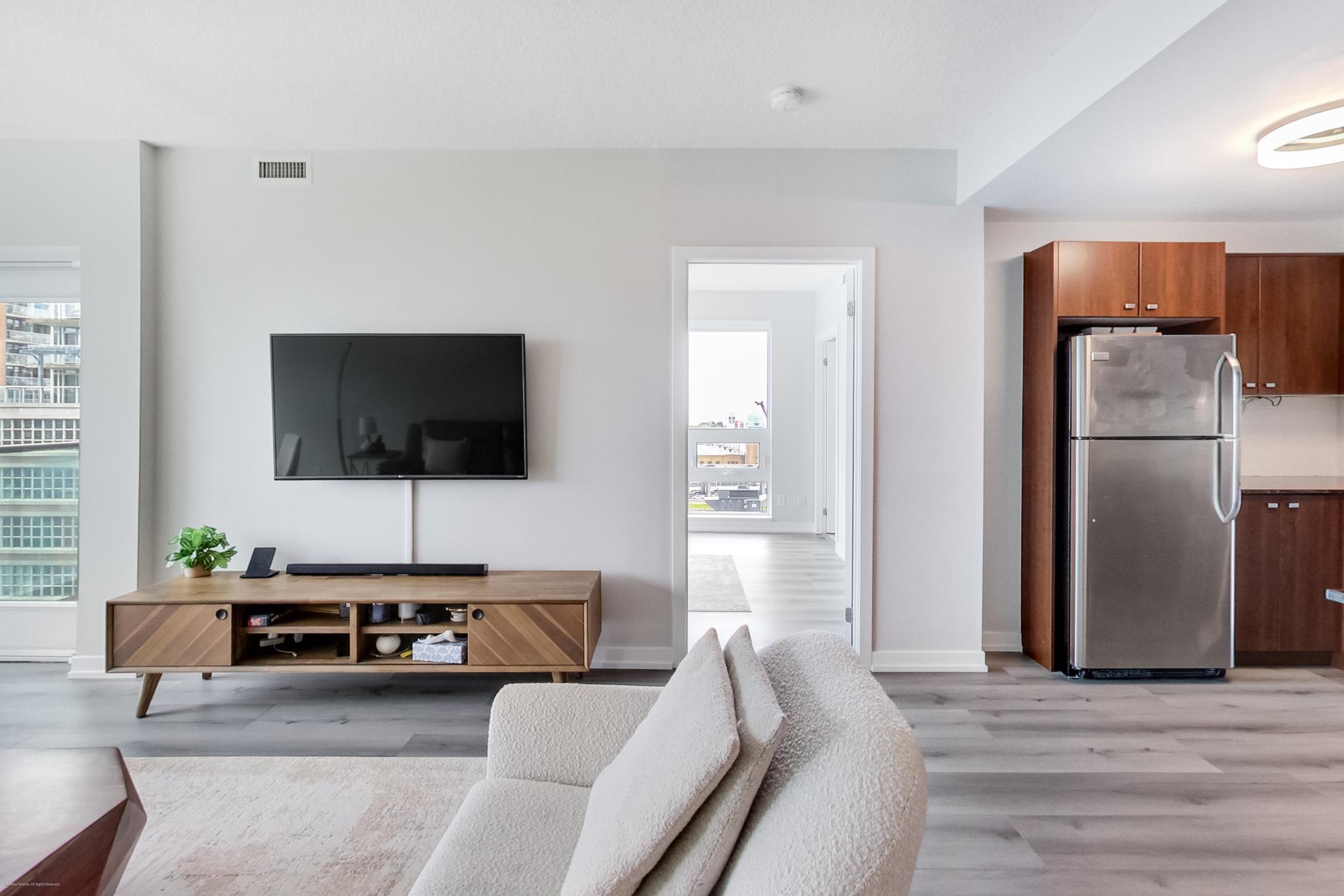 6 | Liberty Village Toronto | Your Online Neighbourhood Guide Living room with a wall-mounted TV above a wooden console, a small plant, and a light gray sofa at the foreground; doorway leads to a kitchen area with stainless steel fridge.
