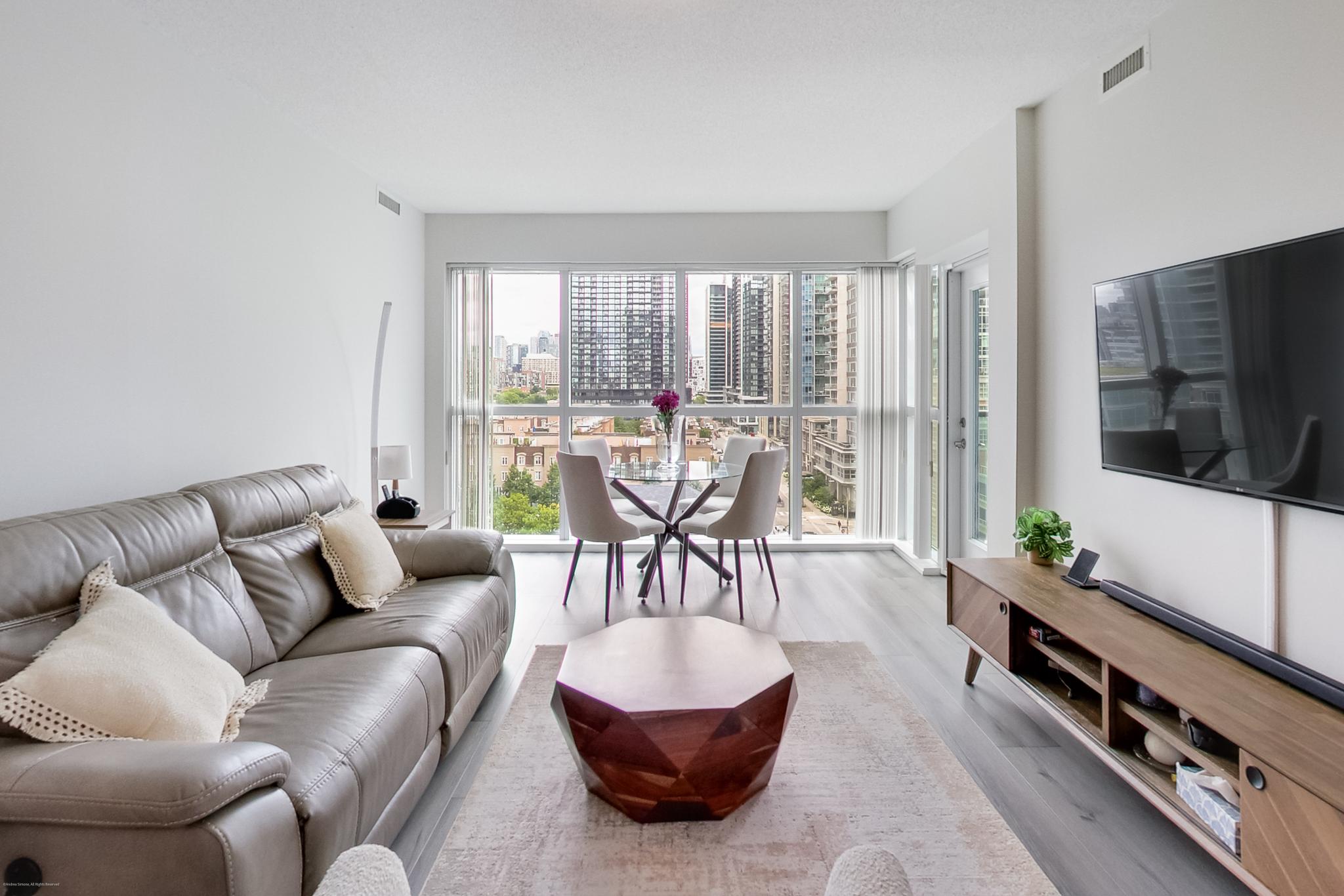 7 | Liberty Village Toronto | Your Online Neighbourhood Guide Bright modern living room with a grey sofa, round wooden coffee table, and dining area by large windows overlooking city buildings.