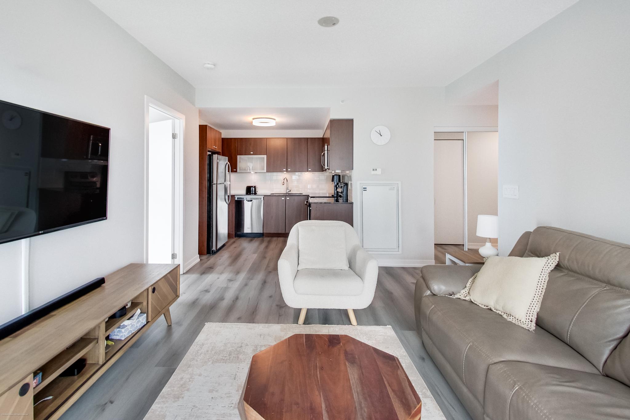 10 | Liberty Village Toronto | Your Online Neighbourhood Guide Open-plan living room with a gray sofa facing a wall-mounted TV and a wood coffee table, kitchen visible in the background.