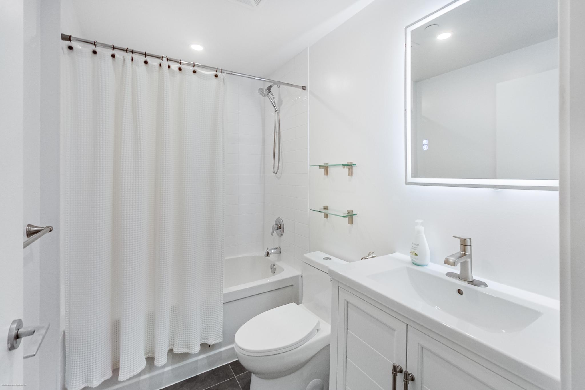 17 | Liberty Village Toronto | Your Online Neighbourhood Guide White bathroom with a tub and shower curtain, wall-mounted showerhead, white tile surround, and a white vanity with a sink and chrome fixtures.