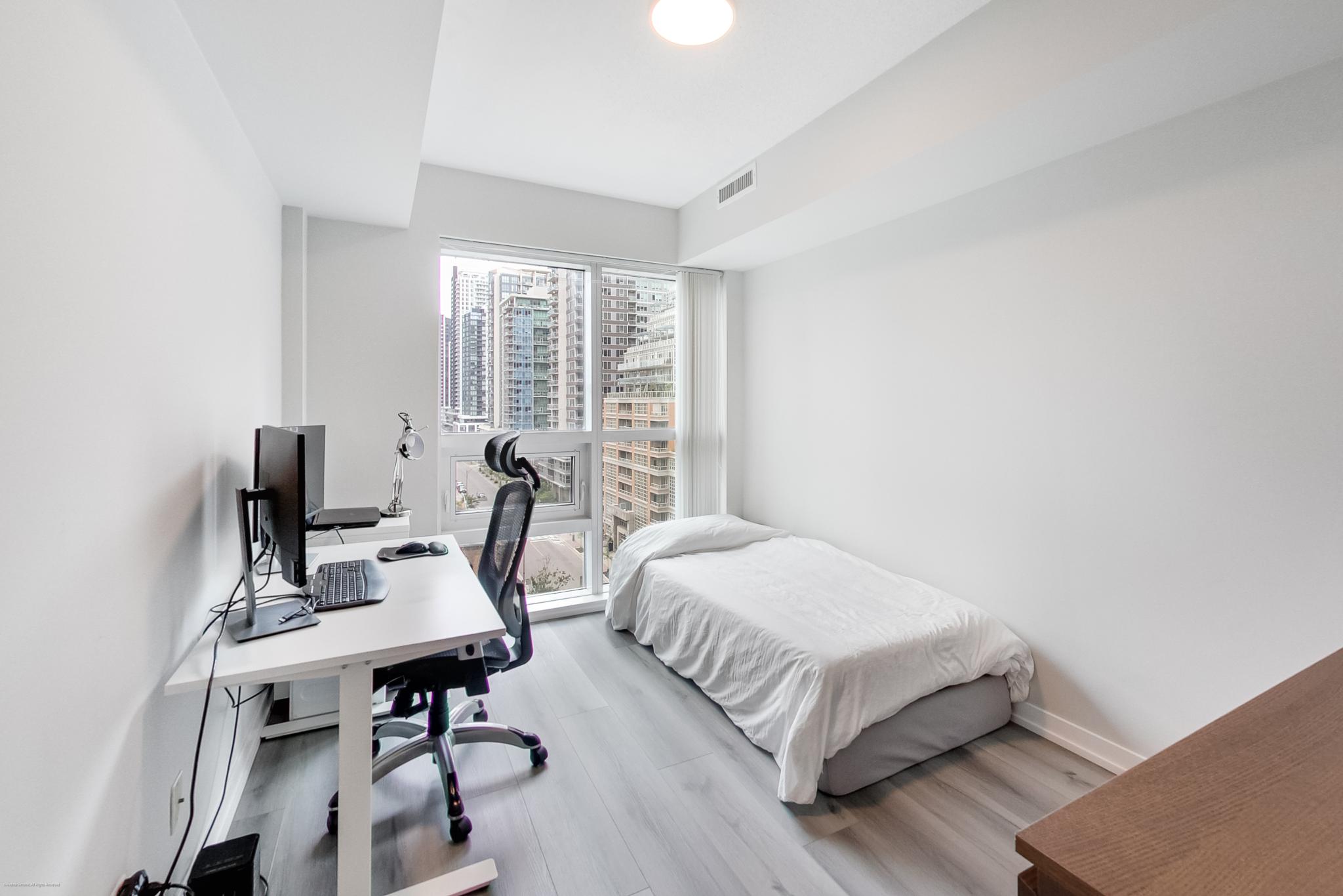 18 | Liberty Village Toronto | Your Online Neighbourhood Guide Small urban bedroom with a white desk and computer chair by a large window, city buildings outside. Bed against the wall on the right.