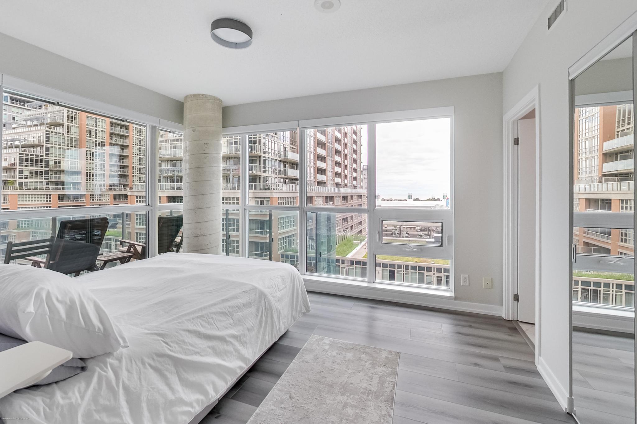 24 | Liberty Village Toronto | Your Online Neighbourhood Guide Bright bedroom with floor-to-ceiling windows showing city high-rise buildings, a white bed in the foreground.