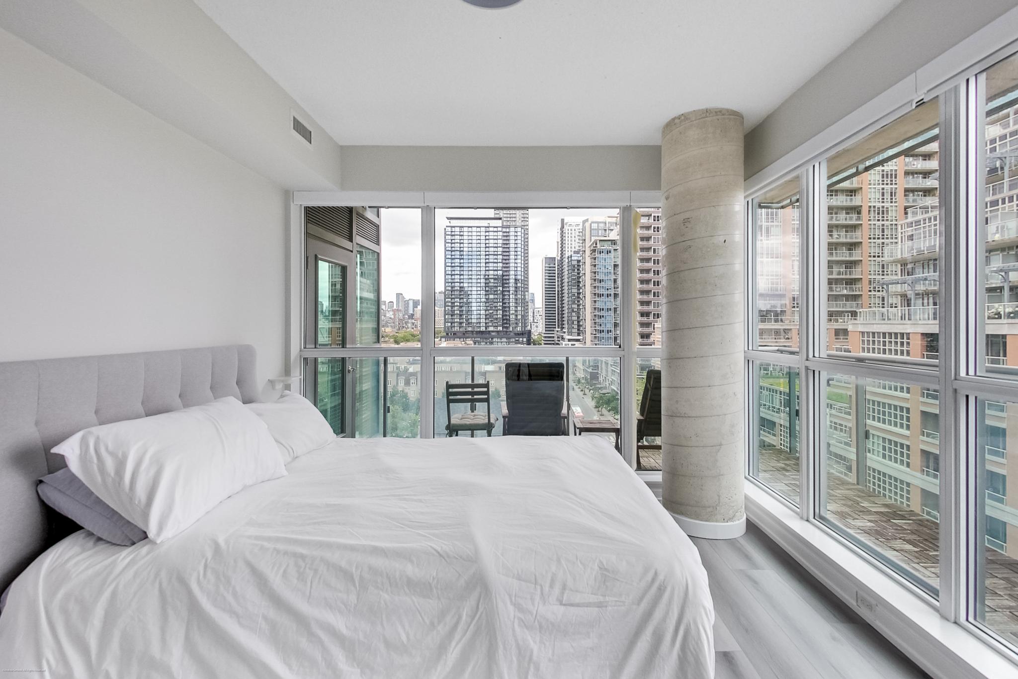 25 | Liberty Village Toronto | Your Online Neighbourhood Guide Bright modern bedroom with floor-to-ceiling windows showing a city skyline, white linens, and a tufted gray headboard.