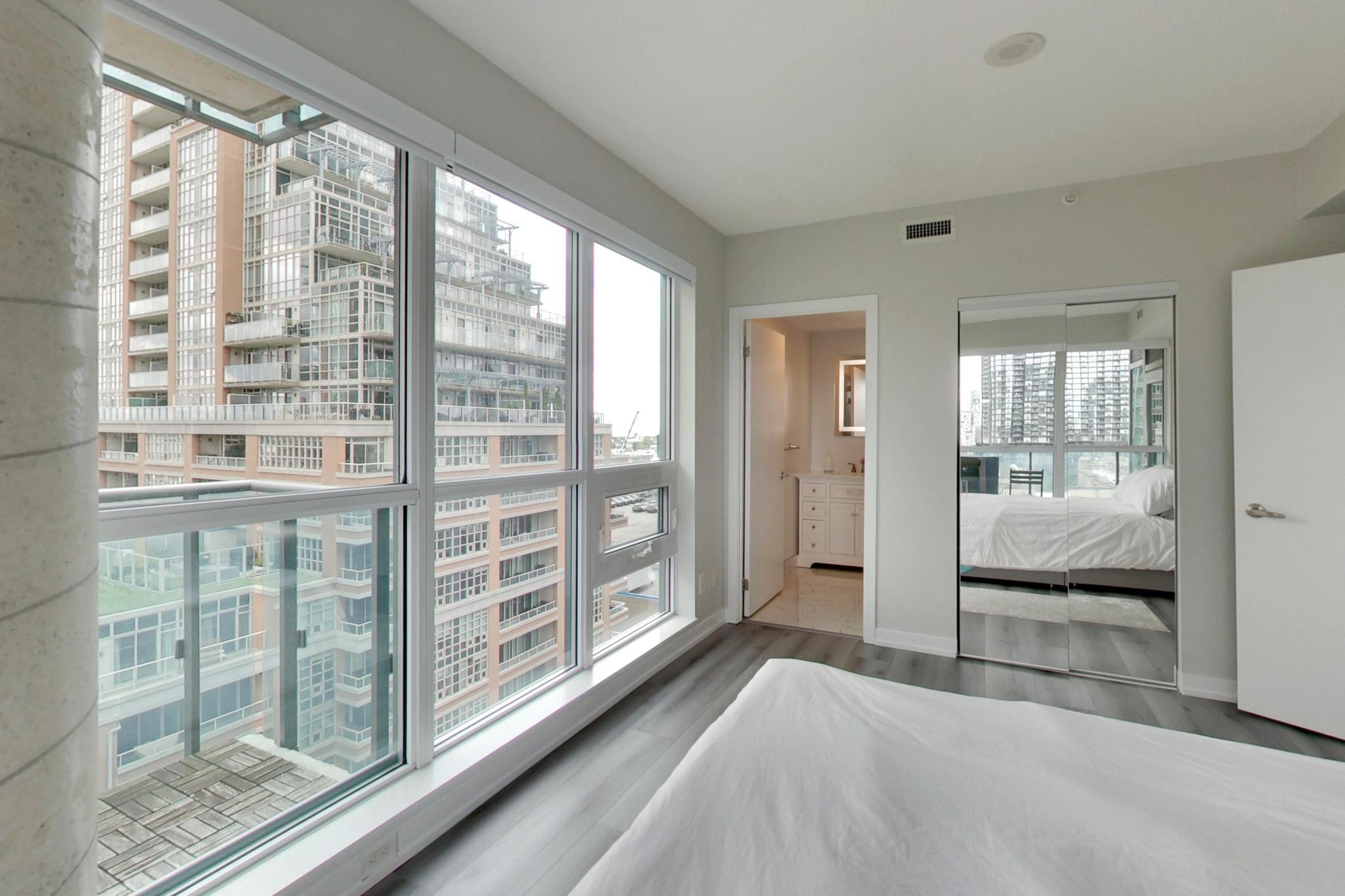 27 | Liberty Village Toronto | Your Online Neighbourhood Guide Bright bedroom with large corner windows overlooking a city high-rise; bed in foreground and a mirrored wardrobe nearby