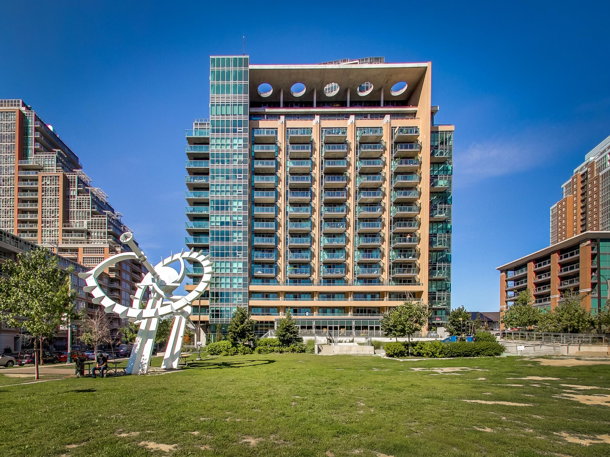 ac_0034 | Liberty Village Toronto | Your Online Neighbourhood Guide Contemporary apartment building with a circular, white sculpture in the foreground and a clear blue sky above. A green lawn and trees frame the scene at a sunny urban park.