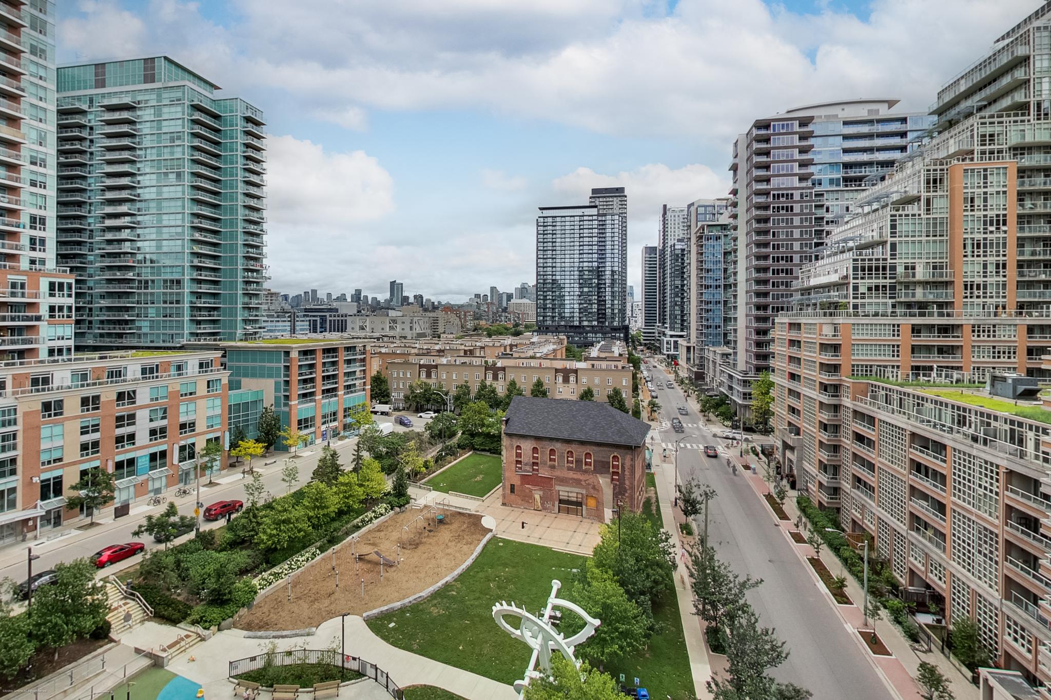 bj_0023 | Liberty Village Toronto | Your Online Neighbourhood Guide Modern city skyline with glass high-rise buildings lining a street and a green park in the foreground.