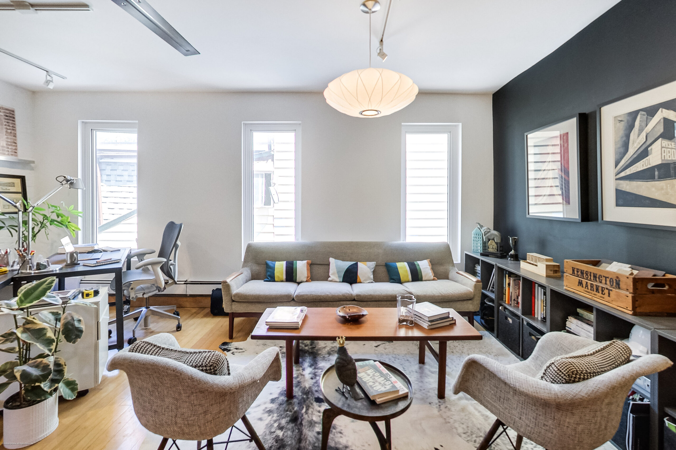 andreasimonecom | Liberty Village Toronto | Your Online Neighbourhood Guide Bright living room with a beige sofa and colorful striped pillows facing a wooden coffee table; three tall windows on the back wall and a dark accent wall with framed art to the right.