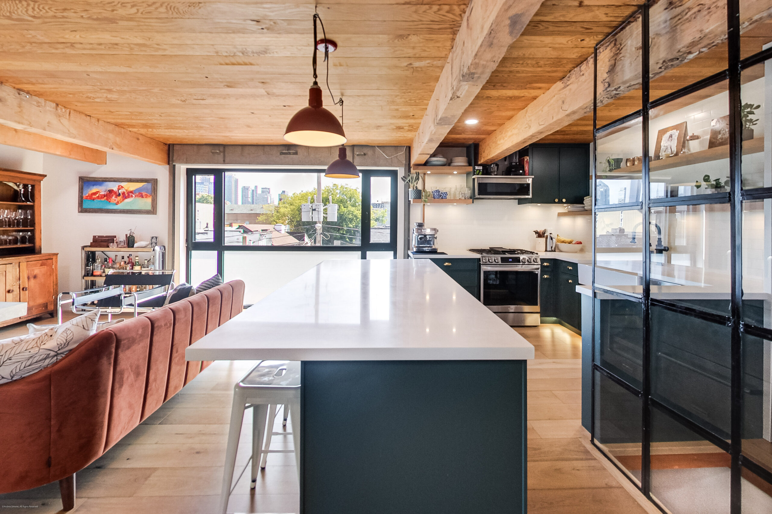 andreasimonecom | Liberty Village Toronto | Your Online Neighbourhood Guide Open-concept kitchen with a long white island, dark green cabinets, and exposed wooden beams; large window shows a cityscape outside.