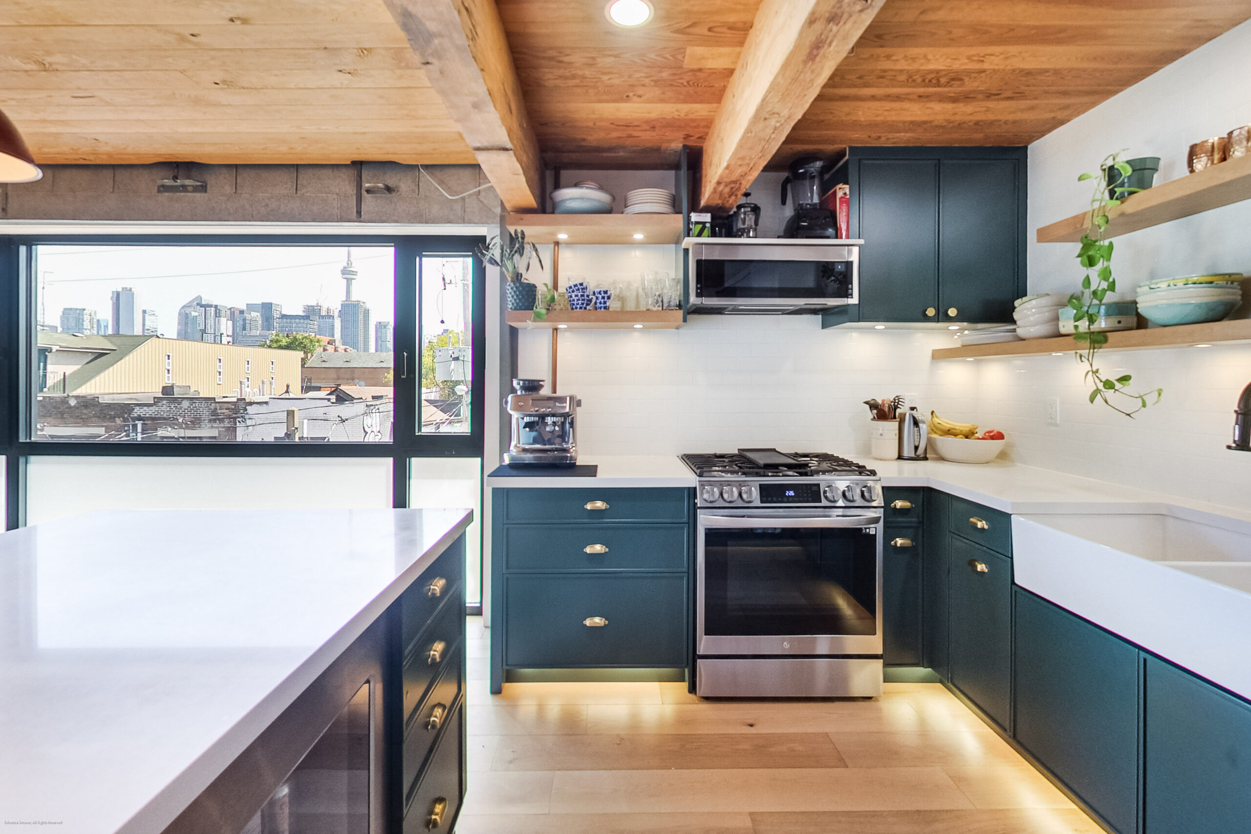andreasimonecom | Liberty Village Toronto | Your Online Neighbourhood Guide Modern kitchen with dark teal cabinets, white countertops, and wooden beam ceiling; city skyline visible through large windows.