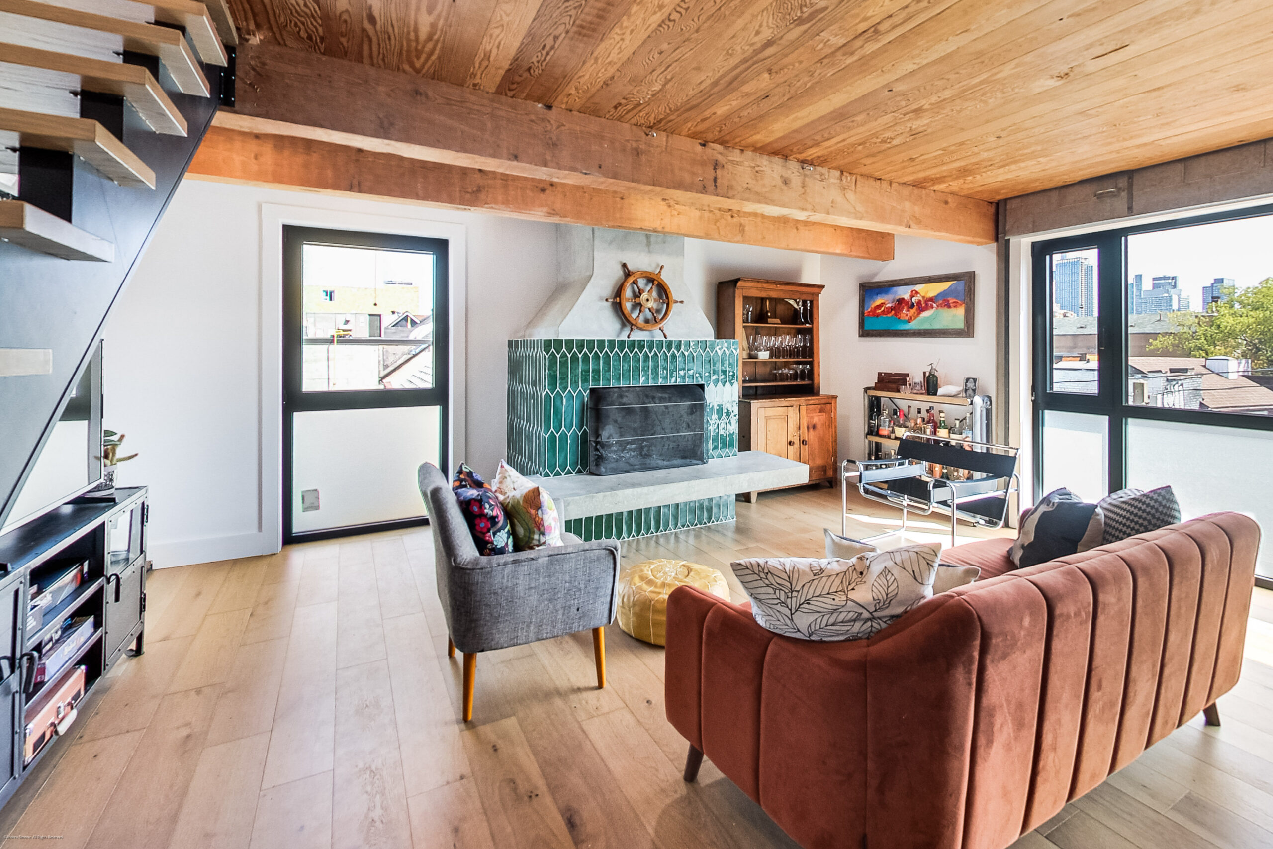 andreasimonecom | Liberty Village Toronto | Your Online Neighbourhood Guide Living room with exposed wooden beams, a teal tiled fireplace, and large windows showing a cityscape outside.
