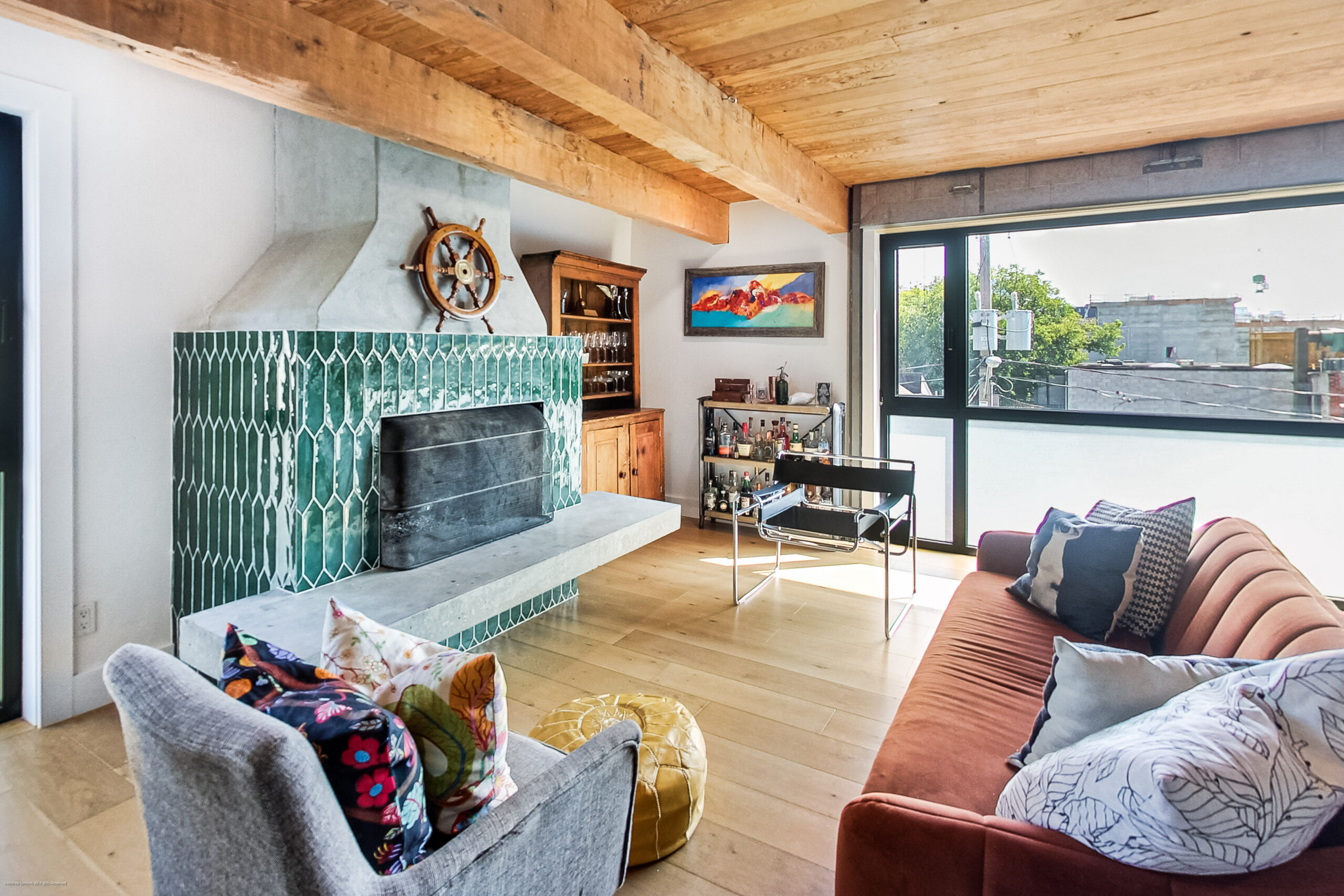 andreasimonecom | Liberty Village Toronto | Your Online Neighbourhood Guide Cozy living room with a green tiled fireplace, wooden beams, and a large window overlooking a cityscape; colorful seating and a bar cart.