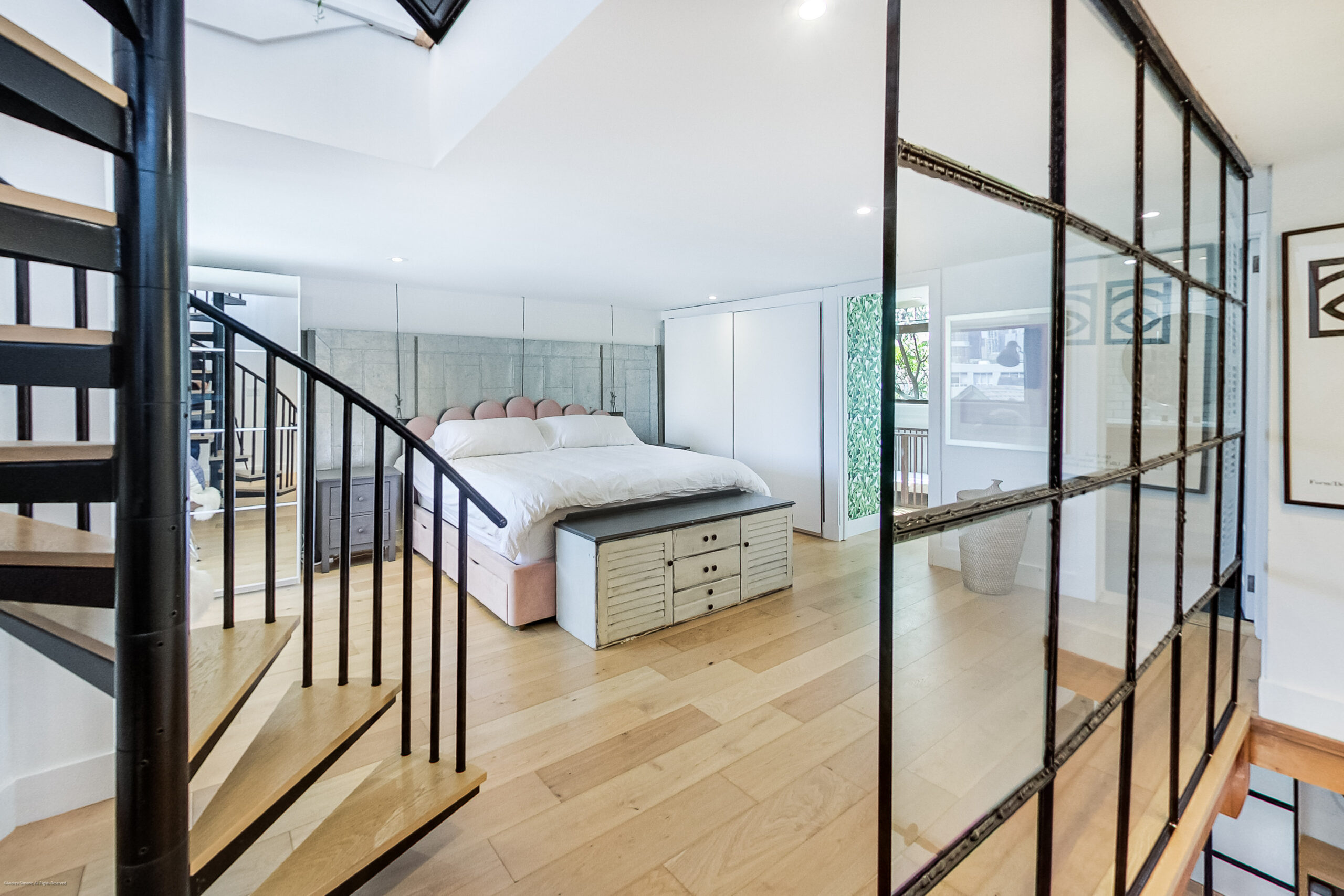 andreasimonecom | Liberty Village Toronto | Your Online Neighbourhood Guide Loft bedroom with a white bed, pink headboard, and light wood floors visible through a black metal-framed glass wall and staircase.