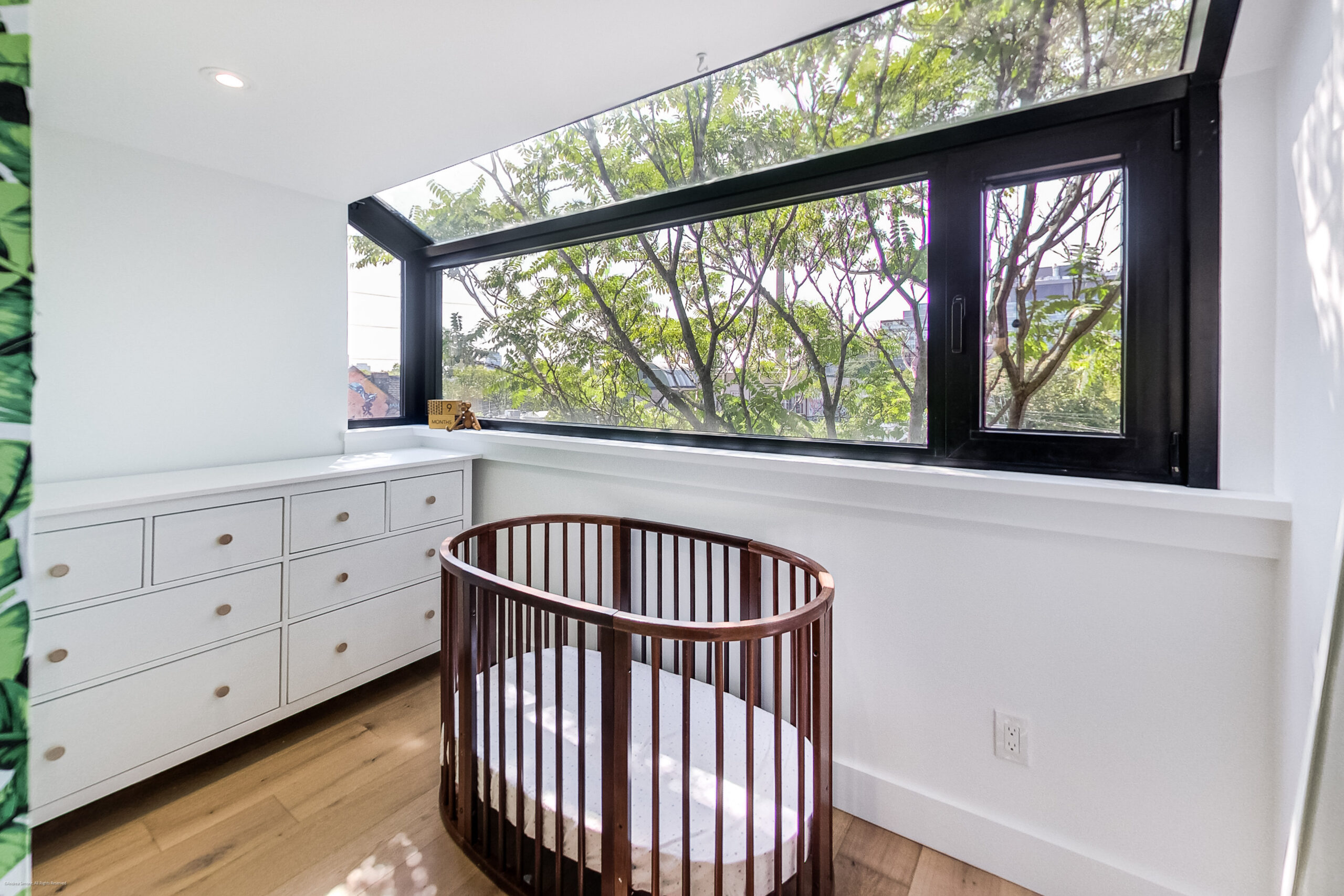 andreasimonecom | Liberty Village Toronto | Your Online Neighbourhood Guide Round wooden baby crib in a bright nursery with white built-in drawers and a large black-framed angled window overlooking trees.