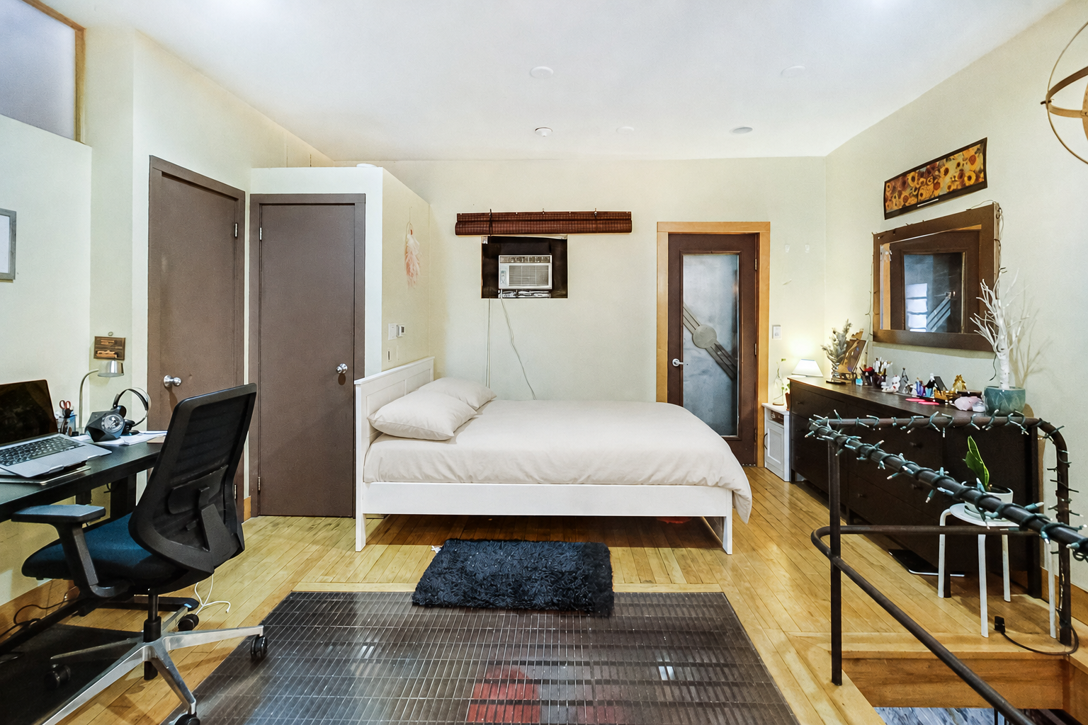 ChatGPT Image 12 ago 2026 10_24_25 | Liberty Village Toronto | Your Online Neighbourhood Guide Bright bedroom with a white-framed bed against a pale wall, a wooden desk and office chair to the left, and a dark dresser with a mirror to the right.