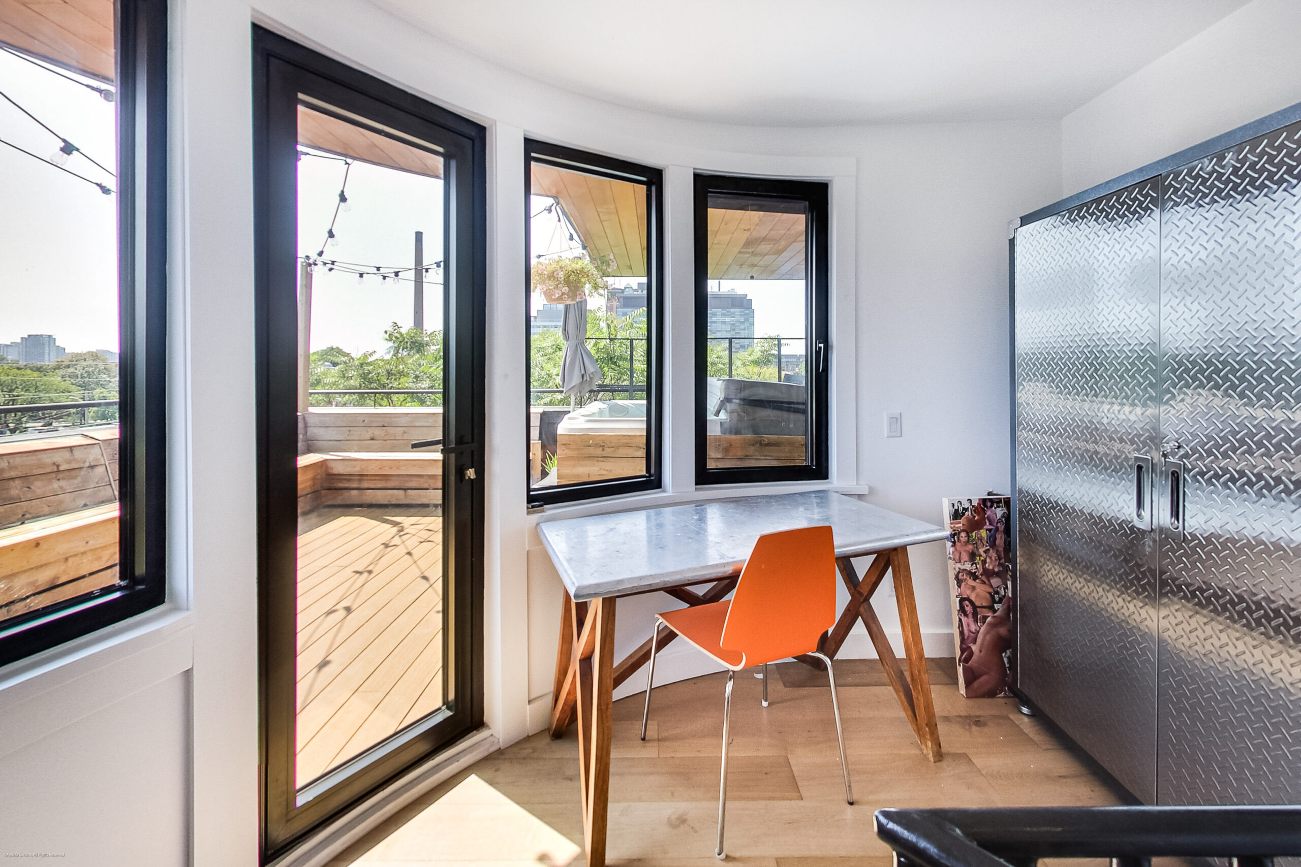 andreasimonecom | Liberty Village Toronto | Your Online Neighbourhood Guide Bright indoor nook with a marble table and orange chairs by large glass sliding doors opening to a sunlit wooden balcony with string lights outside.