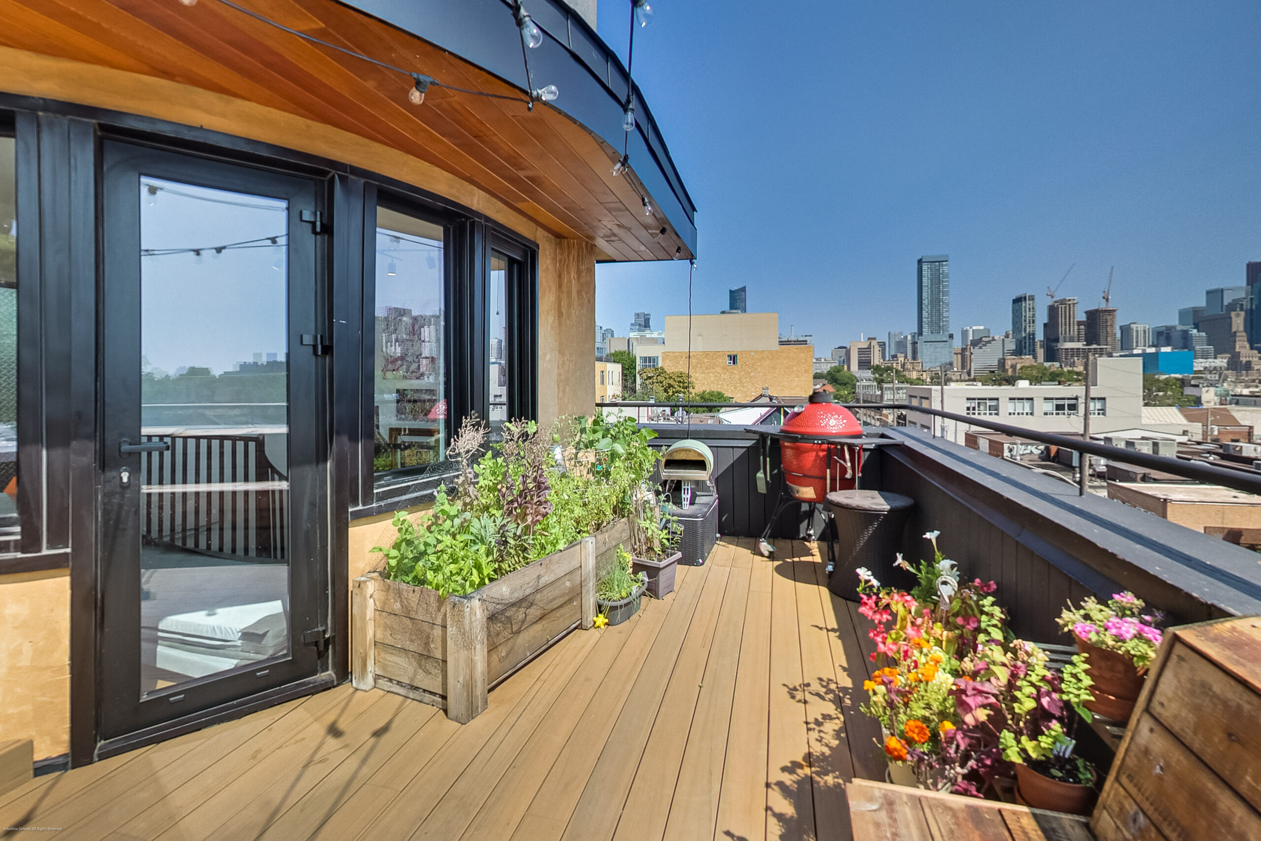 andreasimonecom | Liberty Village Toronto | Your Online Neighbourhood Guide Rooftop deck with wooden flooring, potted plants, black-framed glass doors, and string lights overlooking a city skyline under a clear blue sky.