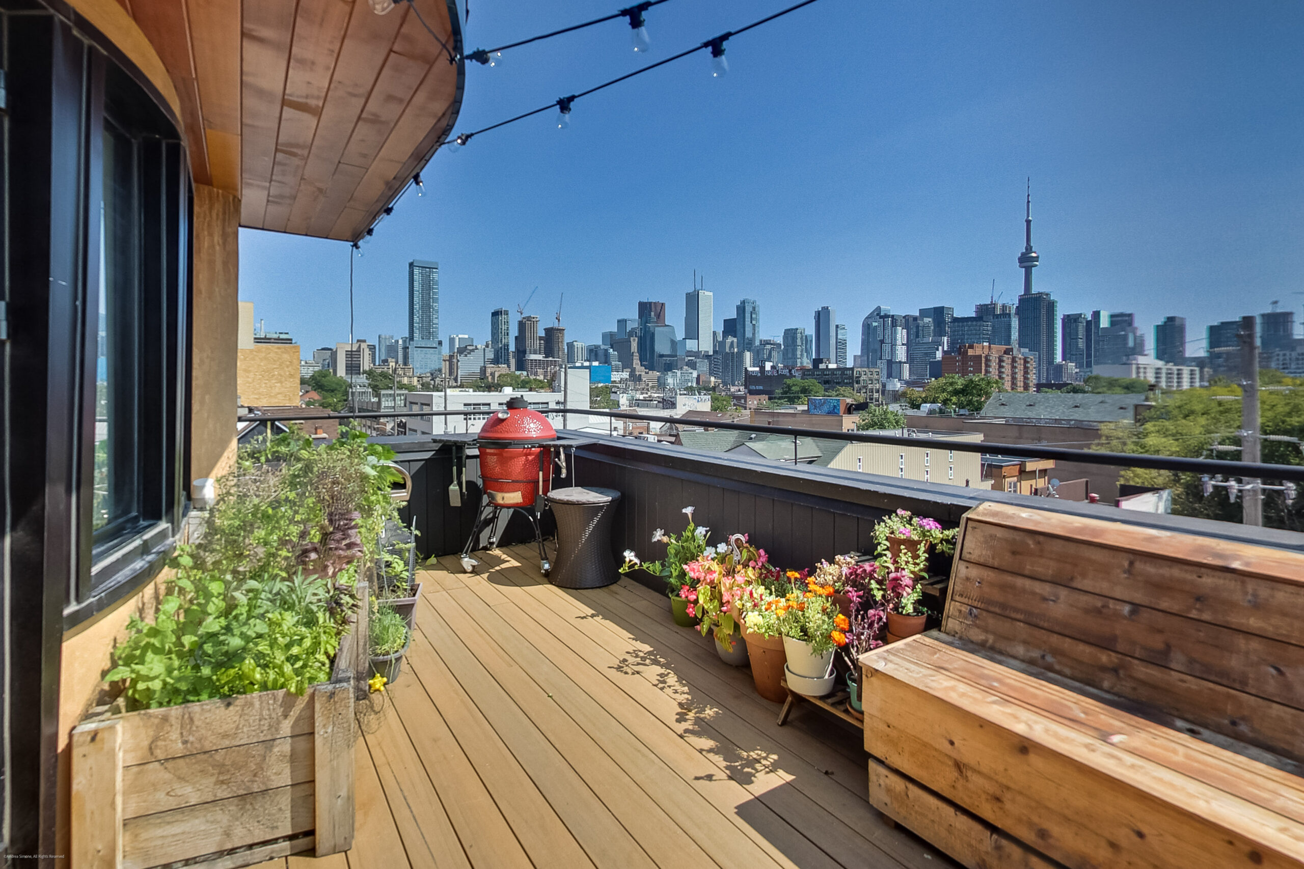 andreasimonecom | Liberty Village Toronto | Your Online Neighbourhood Guide Rooftop terrace with wooden deck, potted flowers, and a red grill; city skyline visible in the distance under a clear blue sky to the right side of the image.