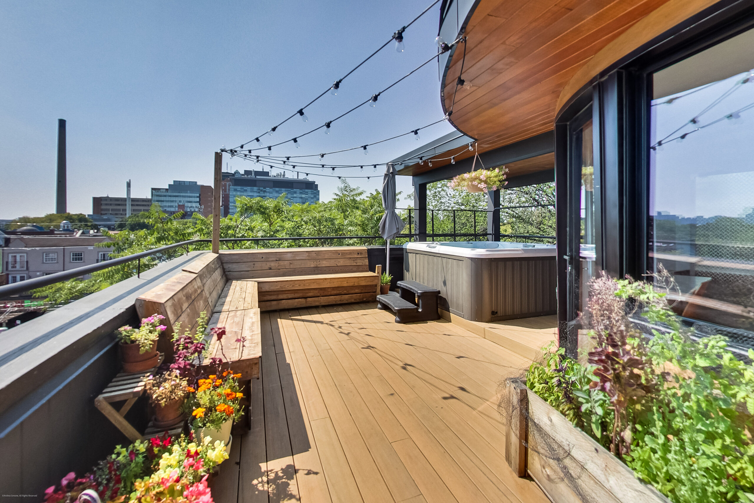 andreasimonecom | Liberty Village Toronto | Your Online Neighbourhood Guide Rooftop deck with string lights, wooden benches, and a hot tub overlooking a city skyline with plants in planters along the edge.